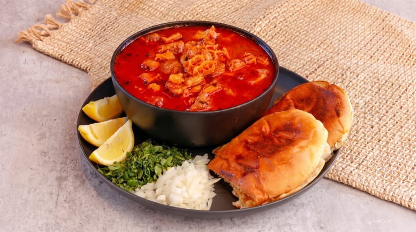 A bowl of red tripe soup with lemon wedges, chopped parsley, onions, and bread on a plate.