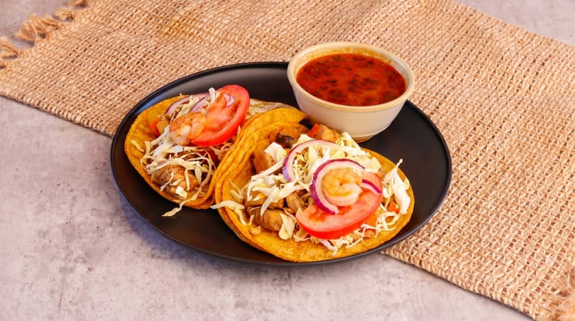 Two shrimp tacos with cabbage, tomato, and red onion, served with a side of red sauce.
