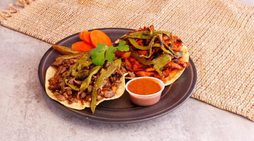 Two tacos with meat, grilled green peppers, and a side of red sauce and carrots.