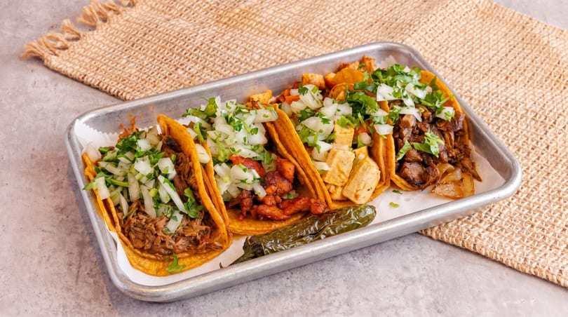 A metal tray holds five tacos with various fillings, topped with diced onions and cilantro.