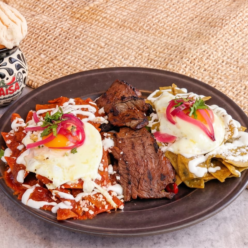 Chilaquiles rojos and verdes with steak and fried eggs, next to a Tacos El Paisa mug.