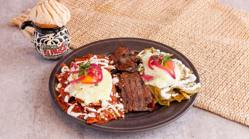 Chilaquiles rojos and verdes with steak and fried eggs, next to a Tacos El Paisa mug.